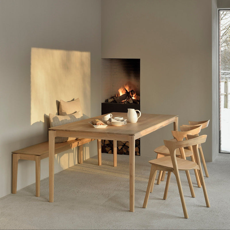 Ethnicraft Bok dining table, bench and chairs in light solid oak beside a lit fireplace