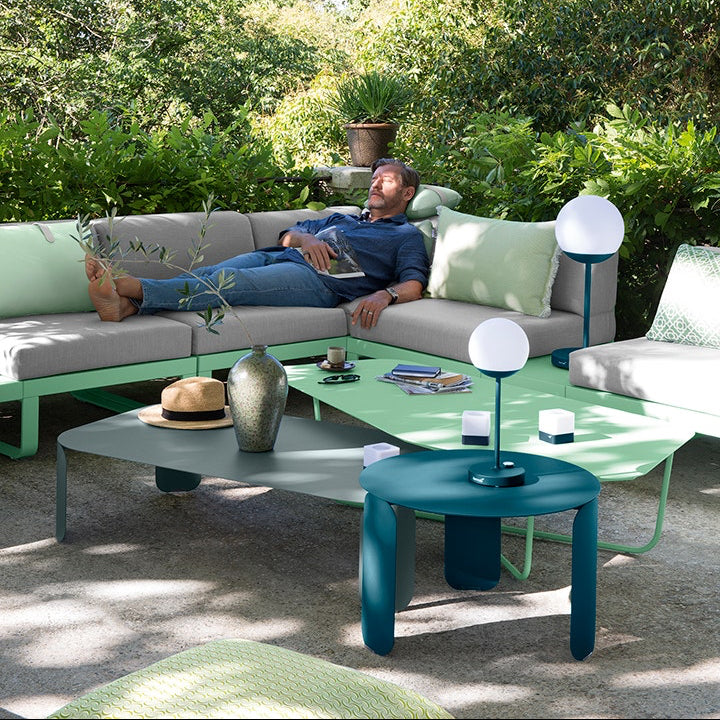 A man reclining on a grey Fermob modular outdoor sofa beside a teal low table and green side table with portable lamps, in a garden