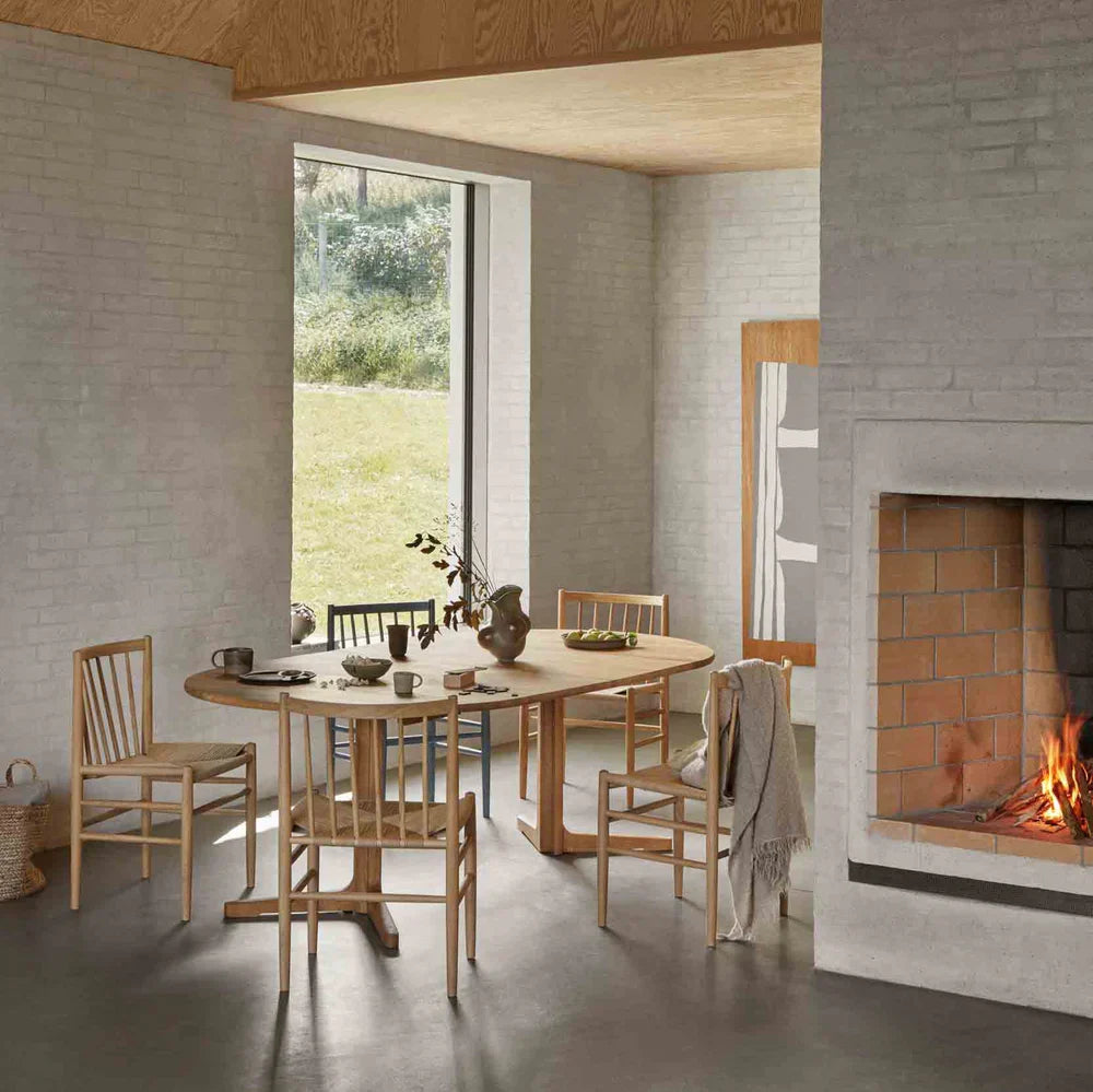 An oak dining table and spindle-back chairs in a whitewashed room with a lit fireplace and a garden view