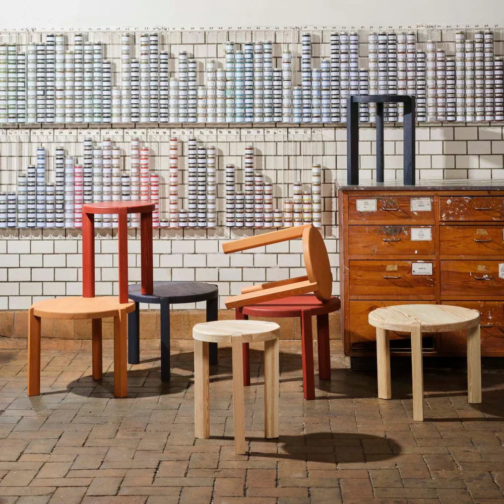 A group of solid wood chairs and stools in natural, red, blue and black finishes beside a vintage cabinet against a tiled wall
