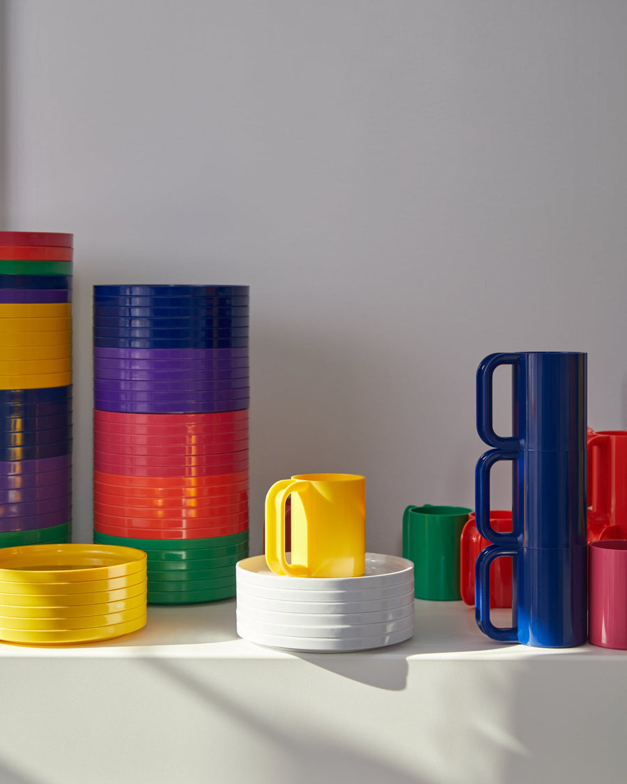 Heller Max dinnerware in many colors: plates stacked in tall columns, white bowls, and mugs stacked into a tower on a white shelf