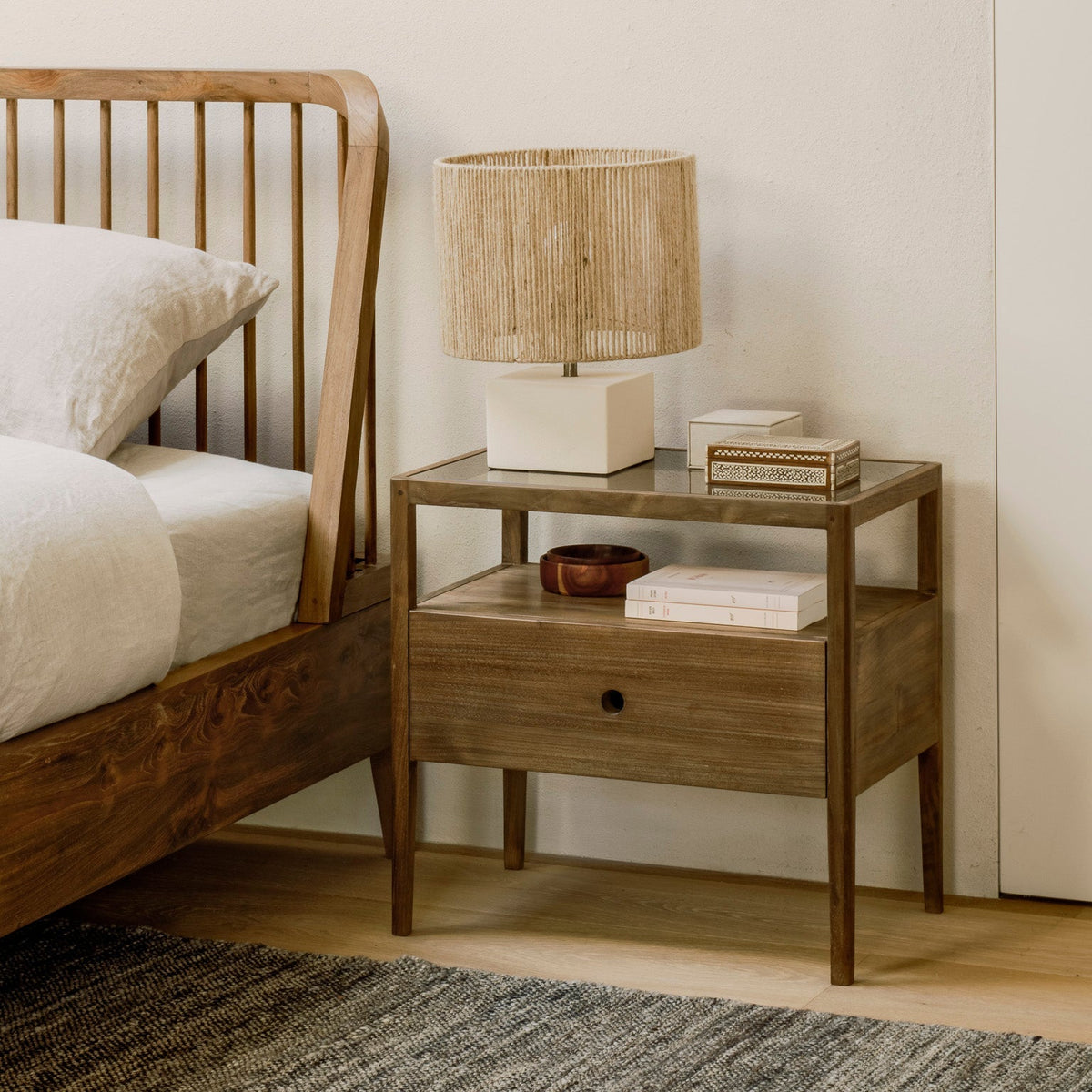 A walnut bedside table with a rattan-shaded lamp beside a spindle bed