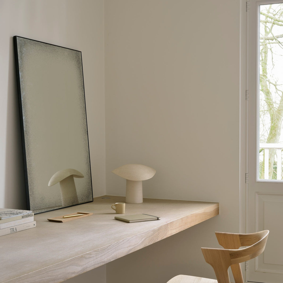 An Ethnicraft Clear wall mirror leaning on a pale oak shelf beside sculptural objects in a sunlit room