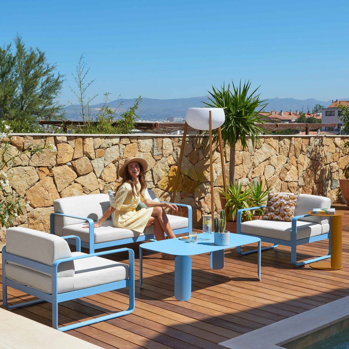 White outdoor lounge chairs and a blue table on a terrace with a stone wall and palms