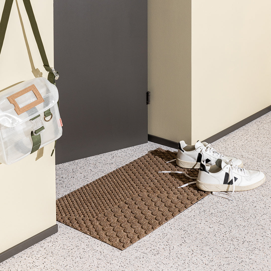 A brown ribbed all-weather doormat in a hallway with sneakers and a canvas bag