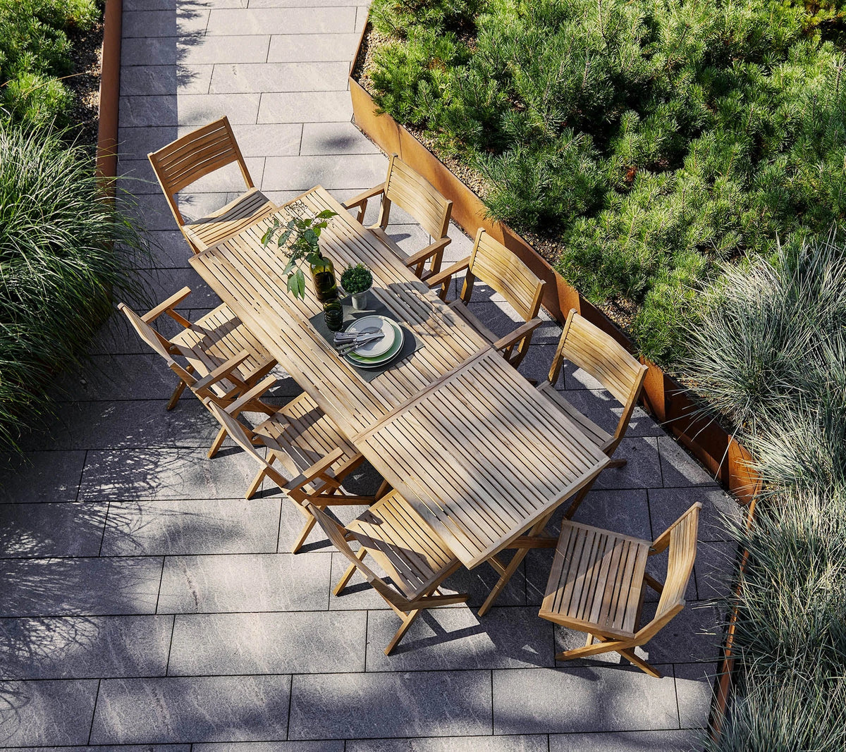 A long teak outdoor dining table with folding chairs on a garden terrace, seen from above