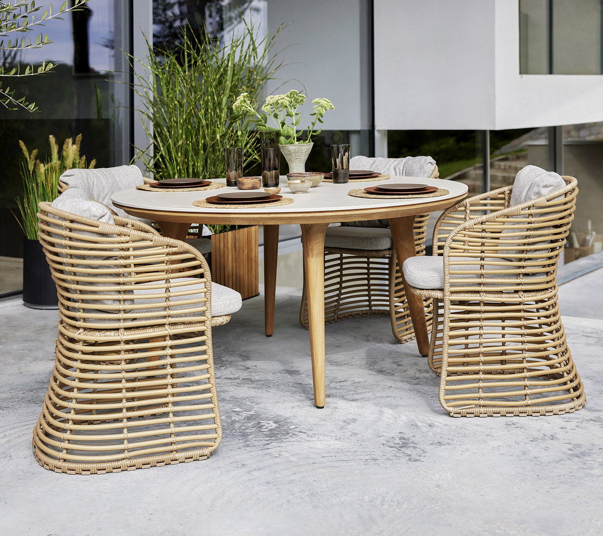 Four Cane-line Basket Dining Chairs in natural Cane-line Weave around a round table, styled in a garden