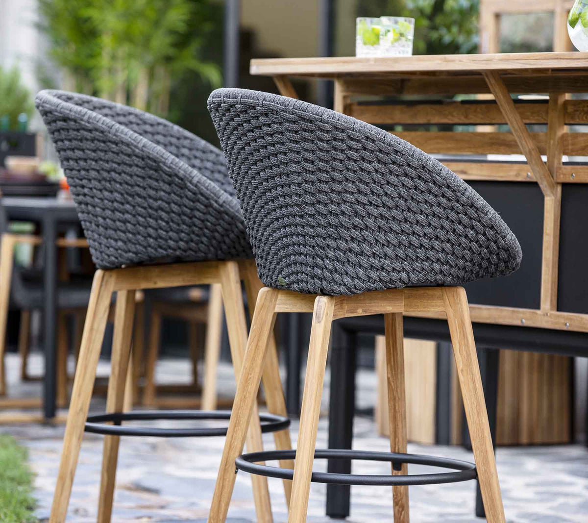 Cane-line Peacock Bar Chairs in Dark Grey lined up at a bar table, styled outdoors, Soft Rope with teak legs