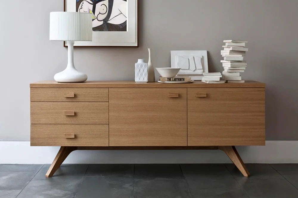 A mid-century modern wooden sideboard credenza with angled legs, styled in a minimalist room with a white table lamp, a framed abstract print, and decorative books.