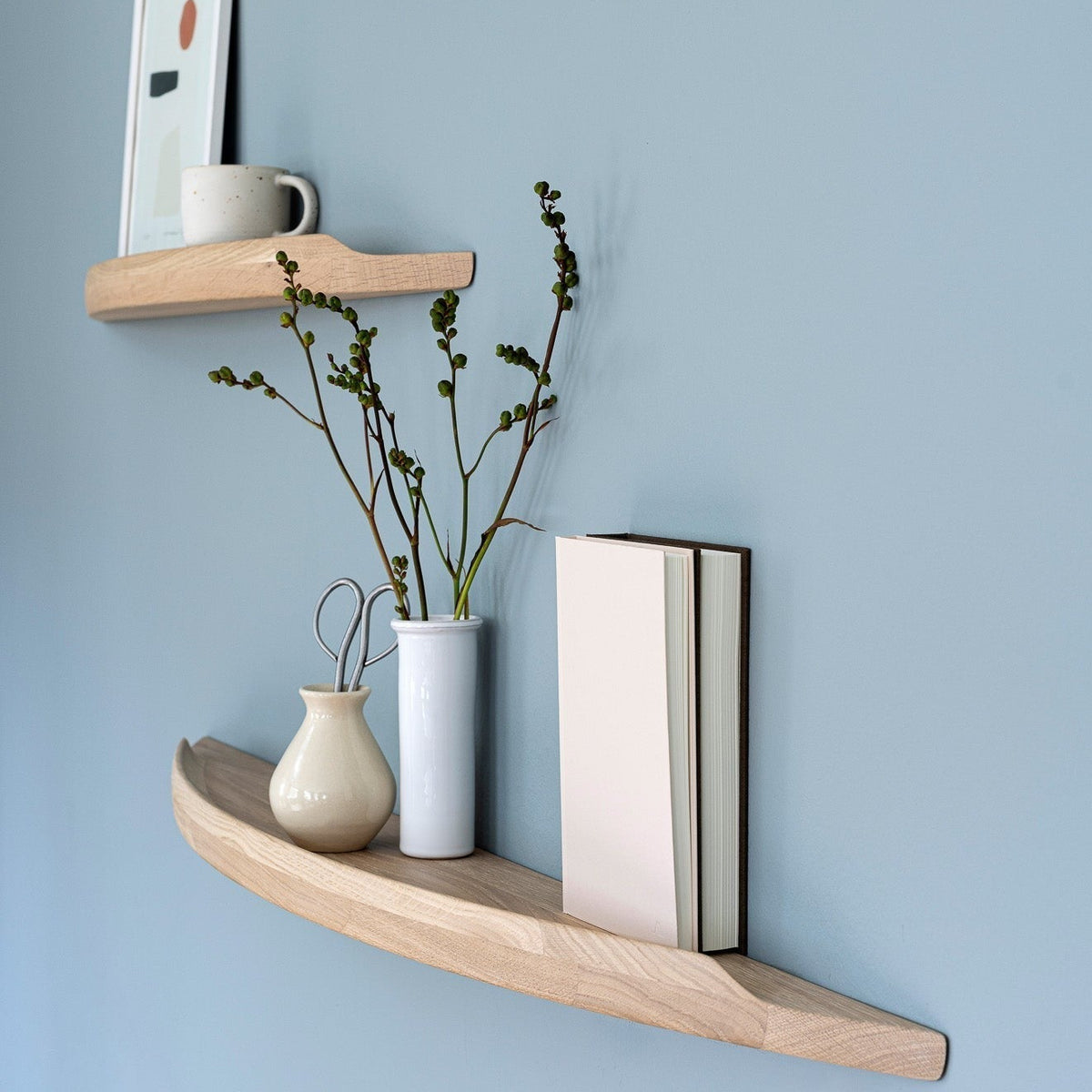 Aakjaer Arcus shelves in white oiled oak holding a print, vases and books on a blue wall
