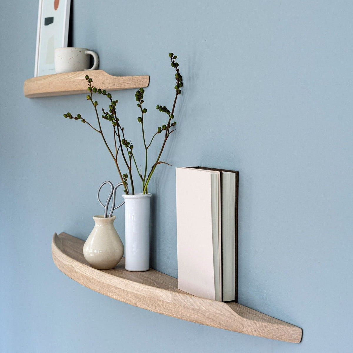 Aakjaer Arcus shelves in white oiled oak holding a print, vases and books on a blue wall