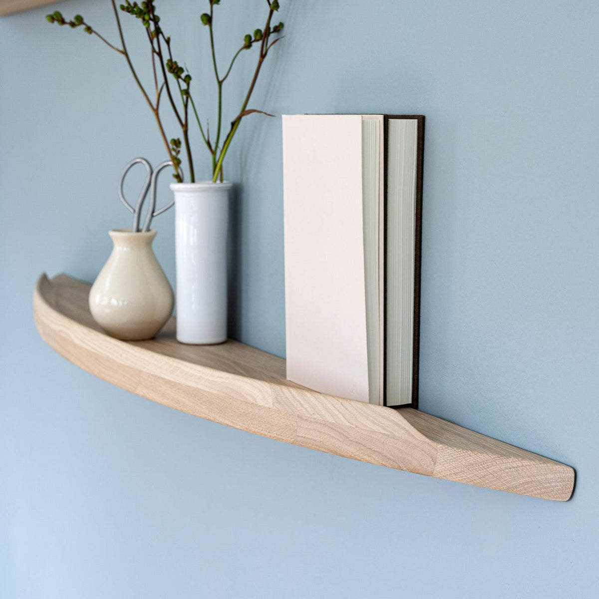 Aakjaer Arcus shelf in white oiled oak with vases and books on a blue living room wall