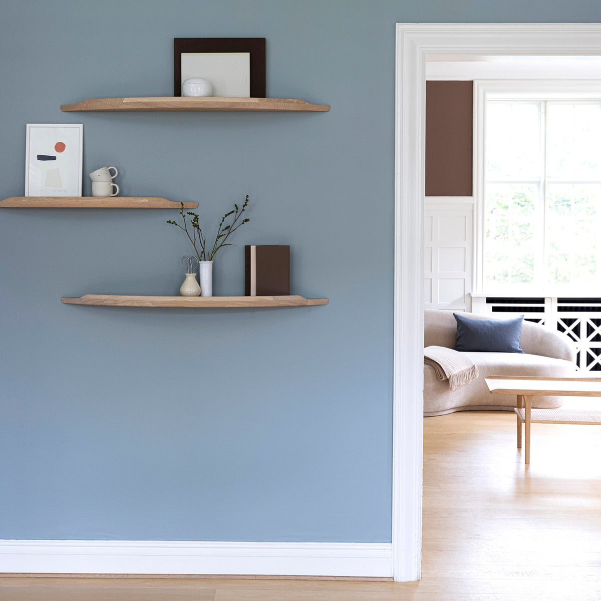 Three Aakjaer Arcus shelves in white oiled oak hung at staggered heights in a blue living room