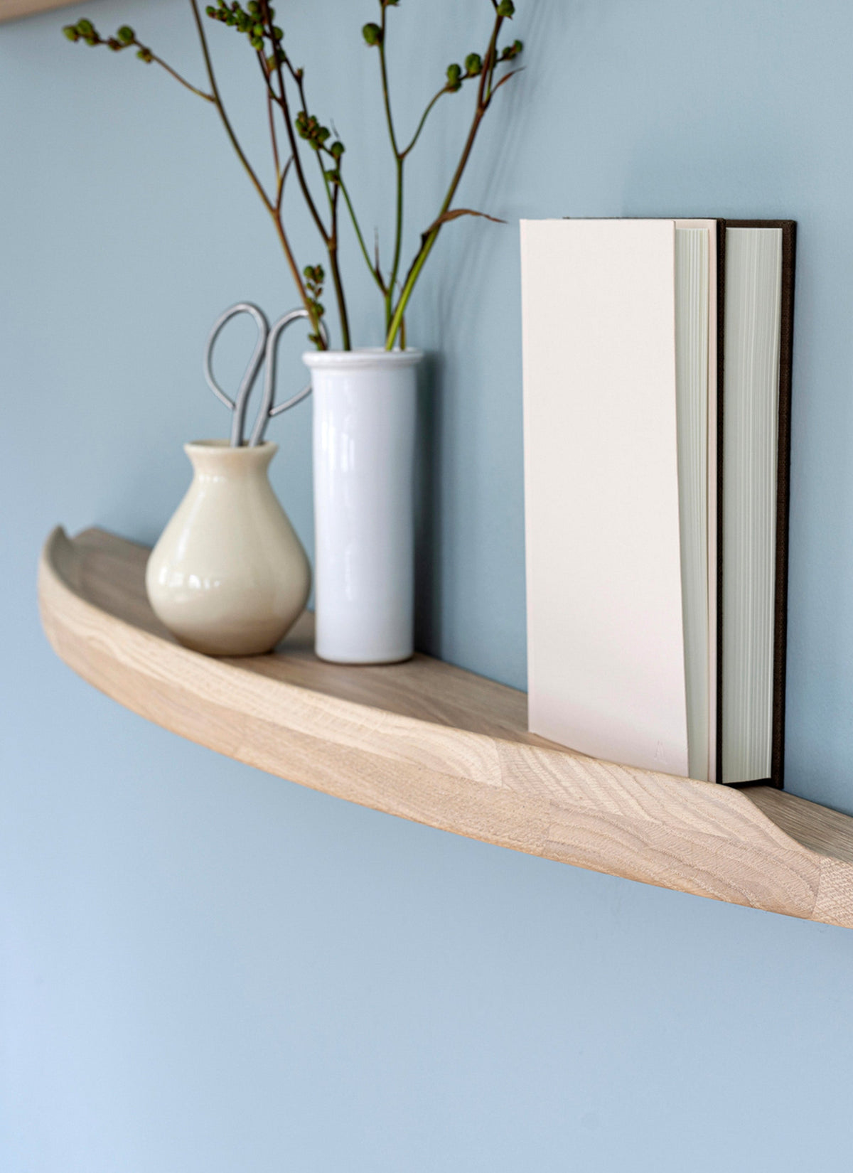 Close-up of an Aakjaer Arcus shelf in white oiled oak holding vases and a book against a blue wall