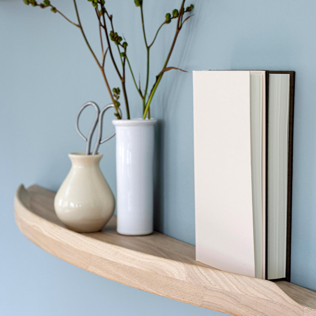 Close-up of an Aakjaer Arcus shelf in white oiled oak holding vases and a book against a blue wall
