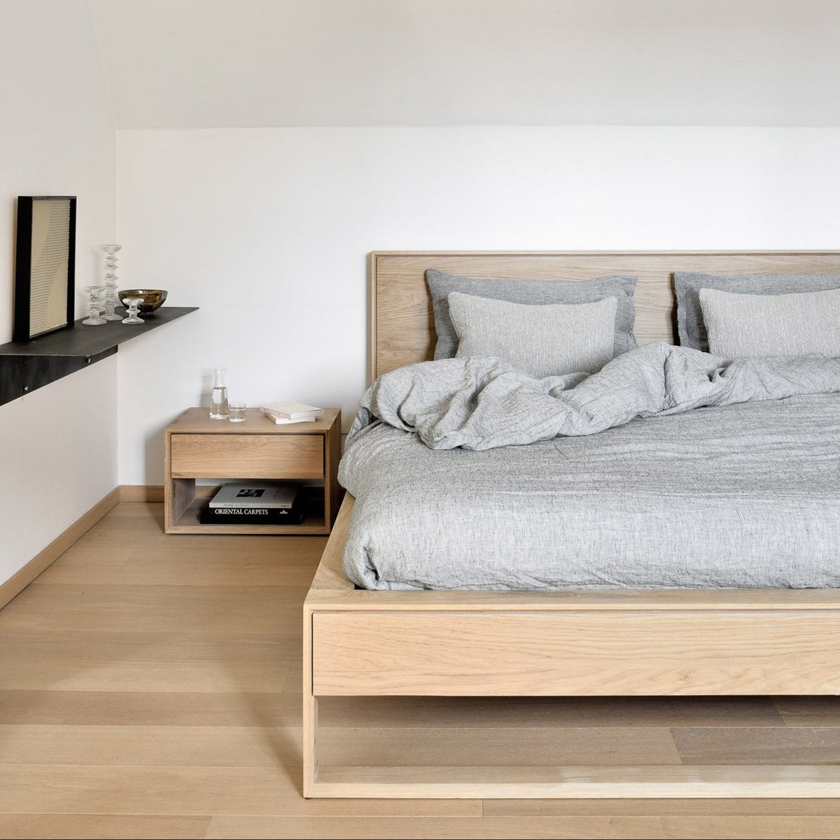Ethnicraft Nordic II bed in light solid oak with gray linen bedding, beside the matching Nordic II bedside table with a drawer and books on its open shelf