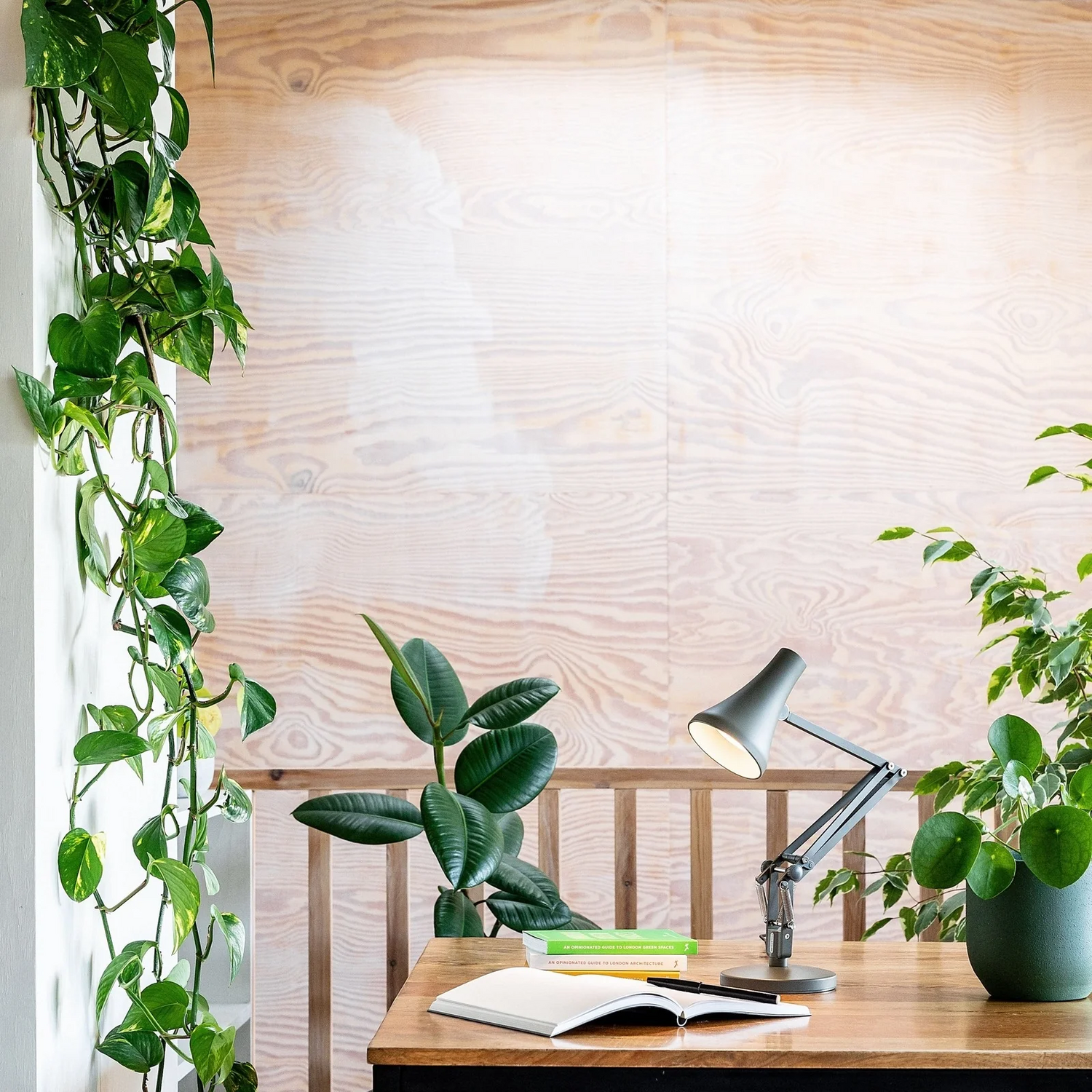 Modern home office setup featuring a wooden desk adorned with a minimalist green adjustable desk lamp, an open book, and green potted plants. A natural wooden backdrop and trailing vines add a refreshing touch of greenery and serenity to the workspace.