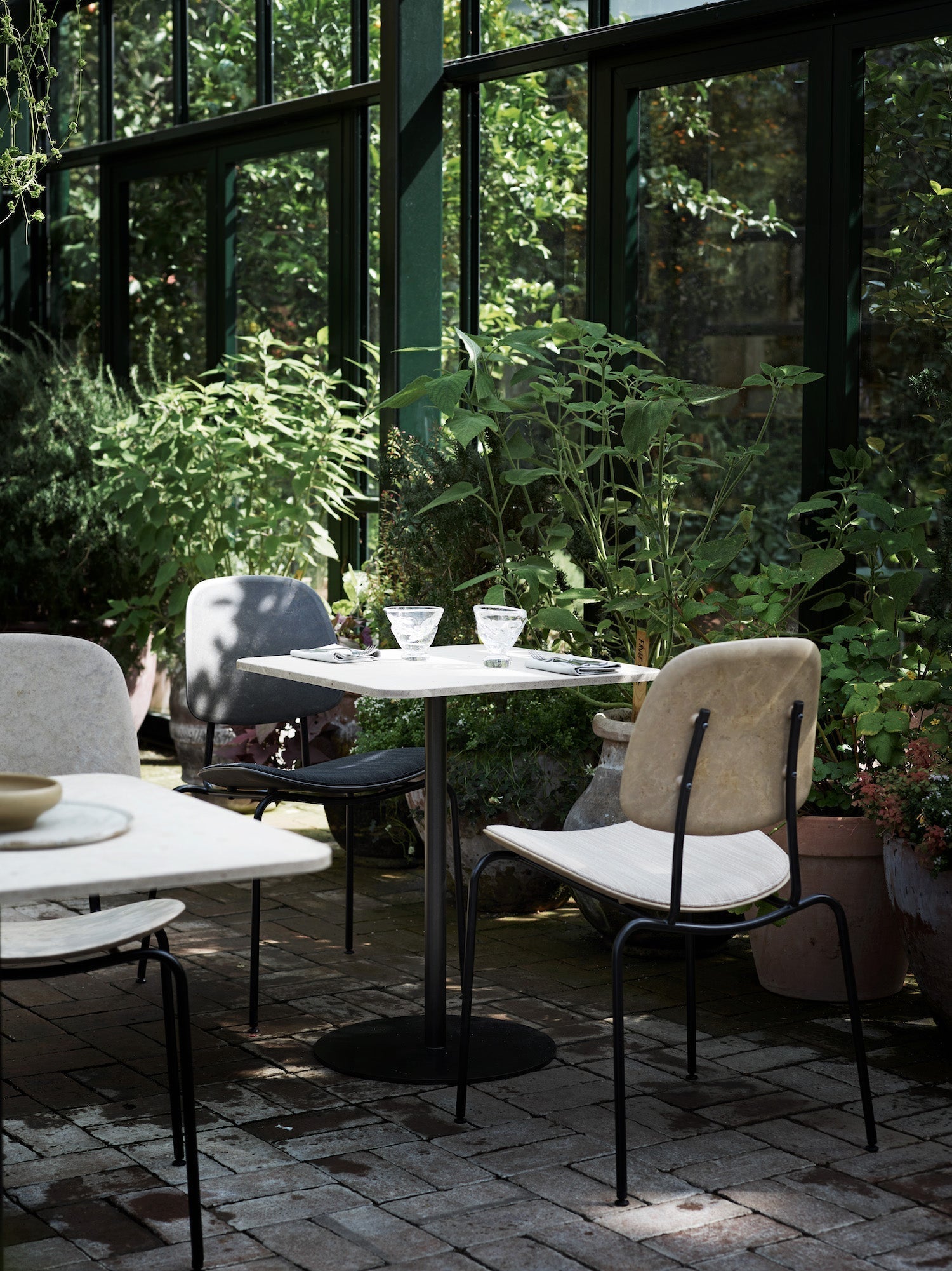 Modern outdoor dining setup featuring sleek black-frame chairs with neutral stone-inspired seats and a minimalist cafe table, surrounded by lush greenery and natural sunlight in a greenhouse-inspired space.