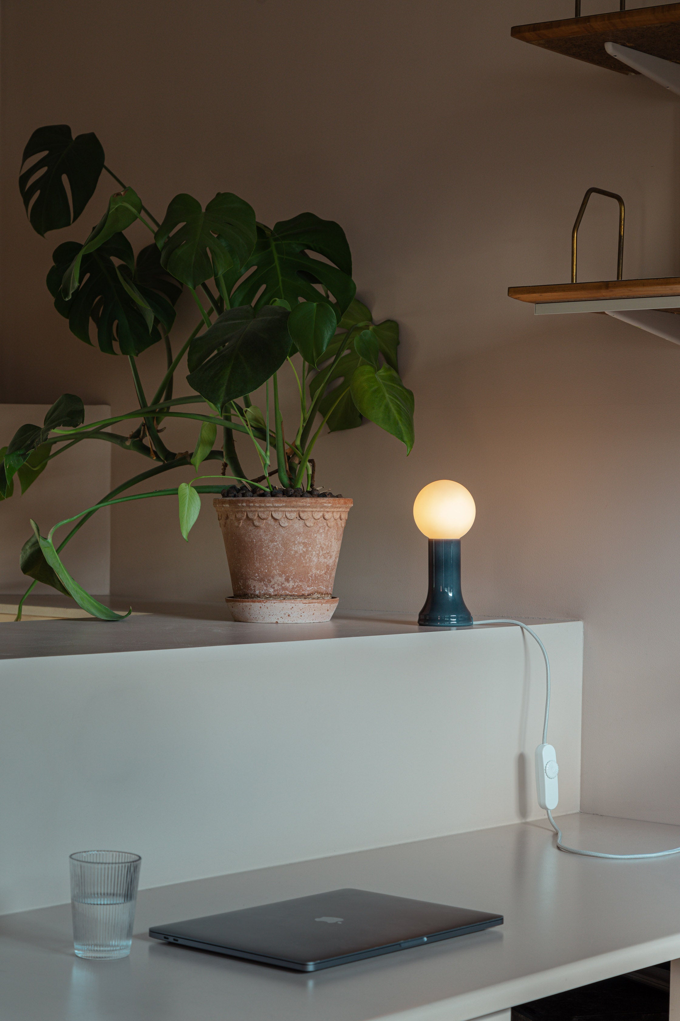 Minimalist bedroom featuring a sleek wall-mounted wooden nightstand with three drawers, a modern sea-green recycled glass table lamp with a glowing spherical bulb, and geometric blue-and-white tiled flooring extending to a visible bathroom.