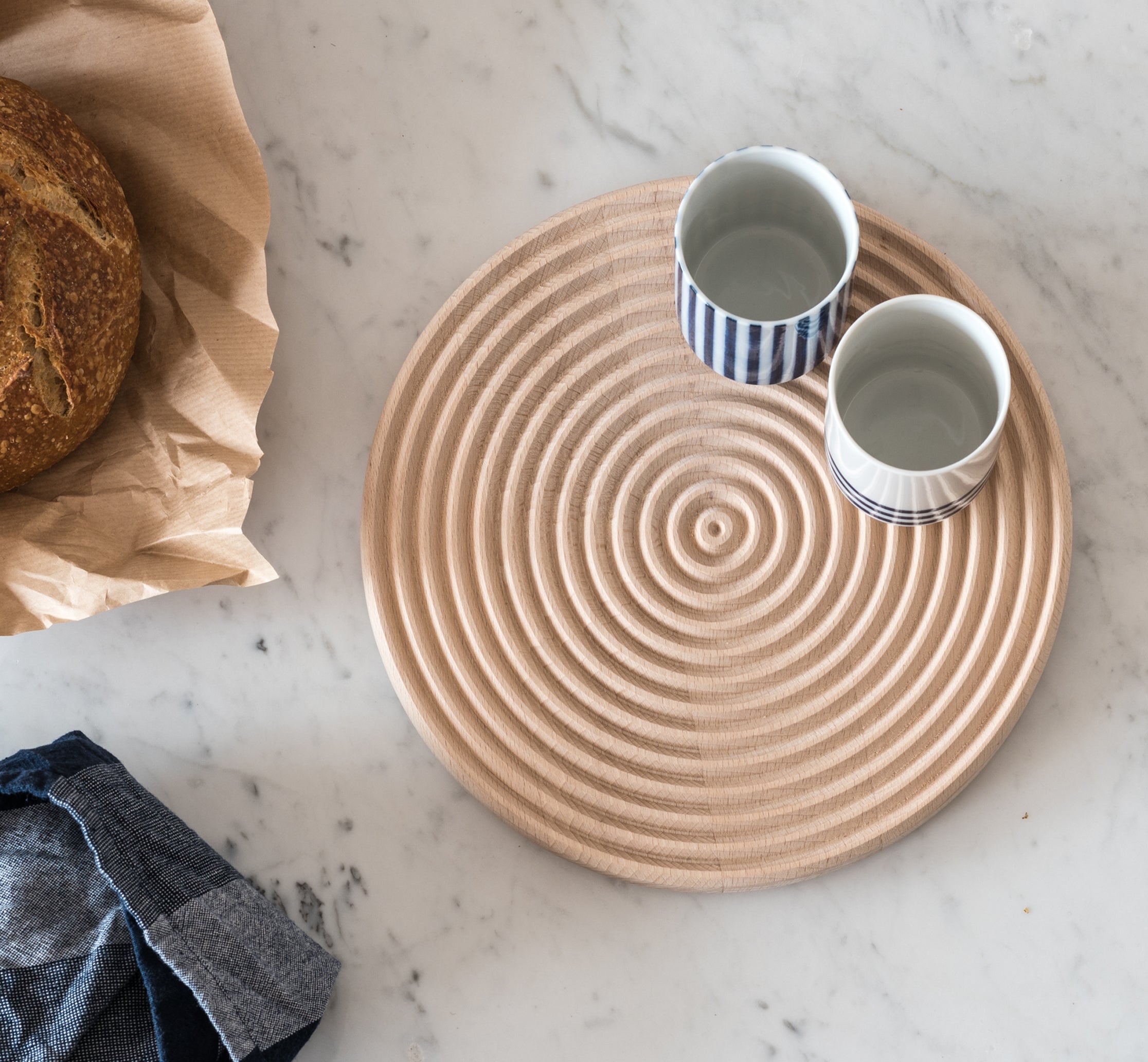 Minimalist tabletop featuring a circular wooden trivet with concentric carved grooves, paired with two ceramic cups in striped patterns, placed on a white marble surface alongside a loaf of artisanal bread wrapped in paper and a folded linen napkin.