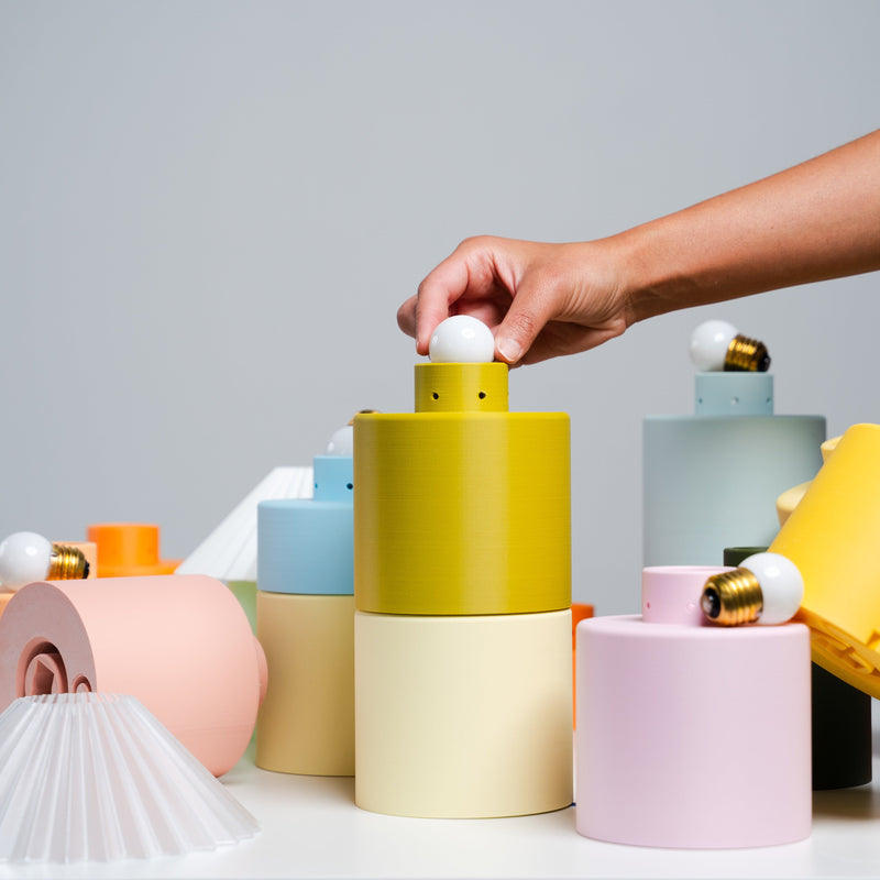 Stacked 3D-printed vessels and pleated lamp shades in yellow, pink, blue and cream, with a hand lifting a lid