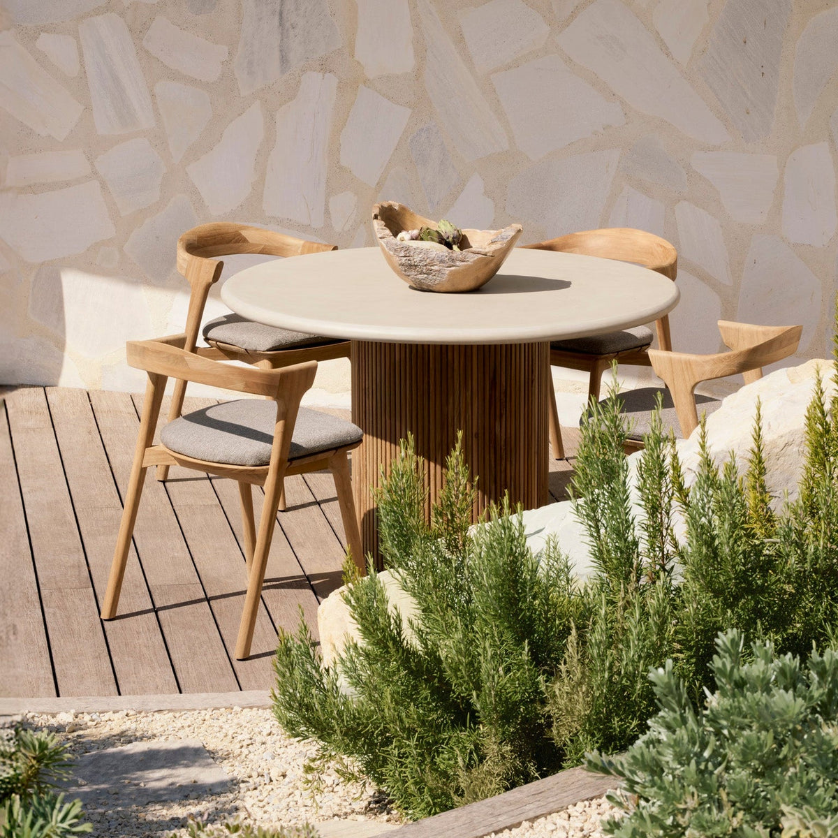 Ethnicraft teak outdoor dining table and chairs on a wooden deck beside a garden