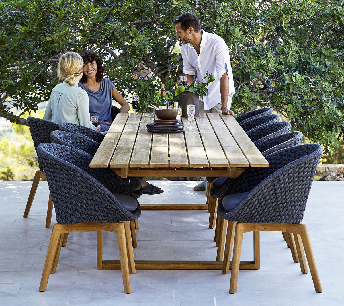Cane-line Endless Dining Table with dining chairs and guests, styled in a garden, solid teak