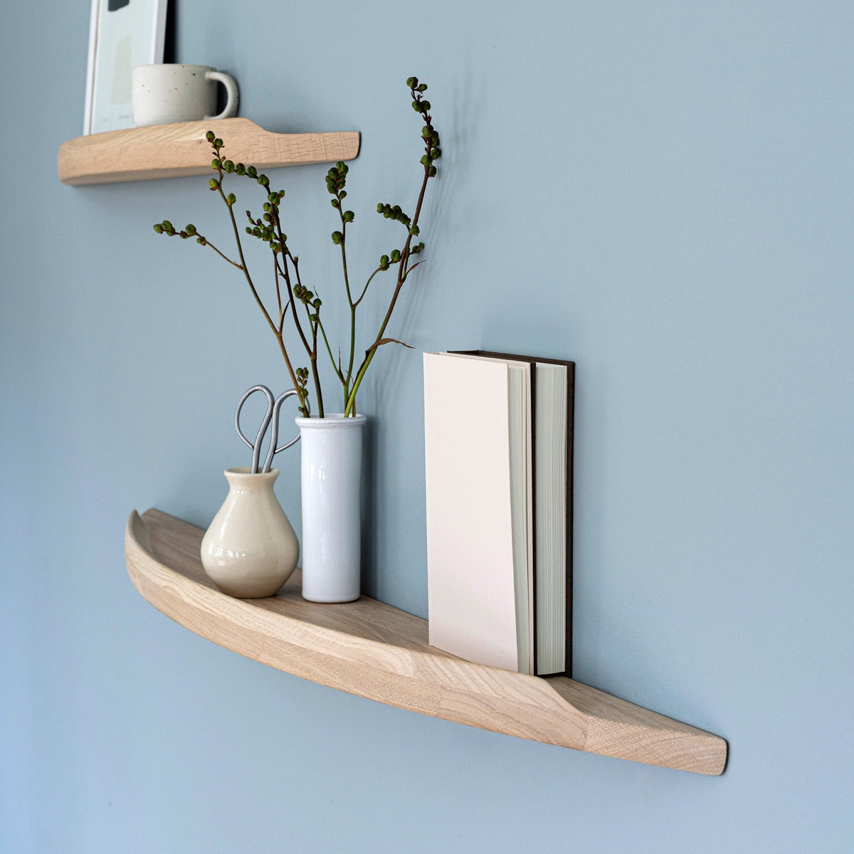 Aakjaer Arcus shelf in white oiled oak with a vase, green stems and books on a blue wall