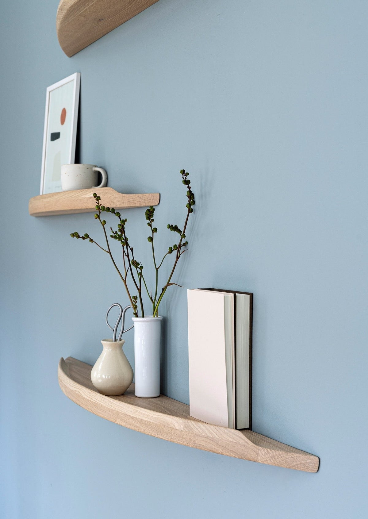 Aakjaer Arcus shelves in white oiled oak holding a print, vases and books on a blue wall
