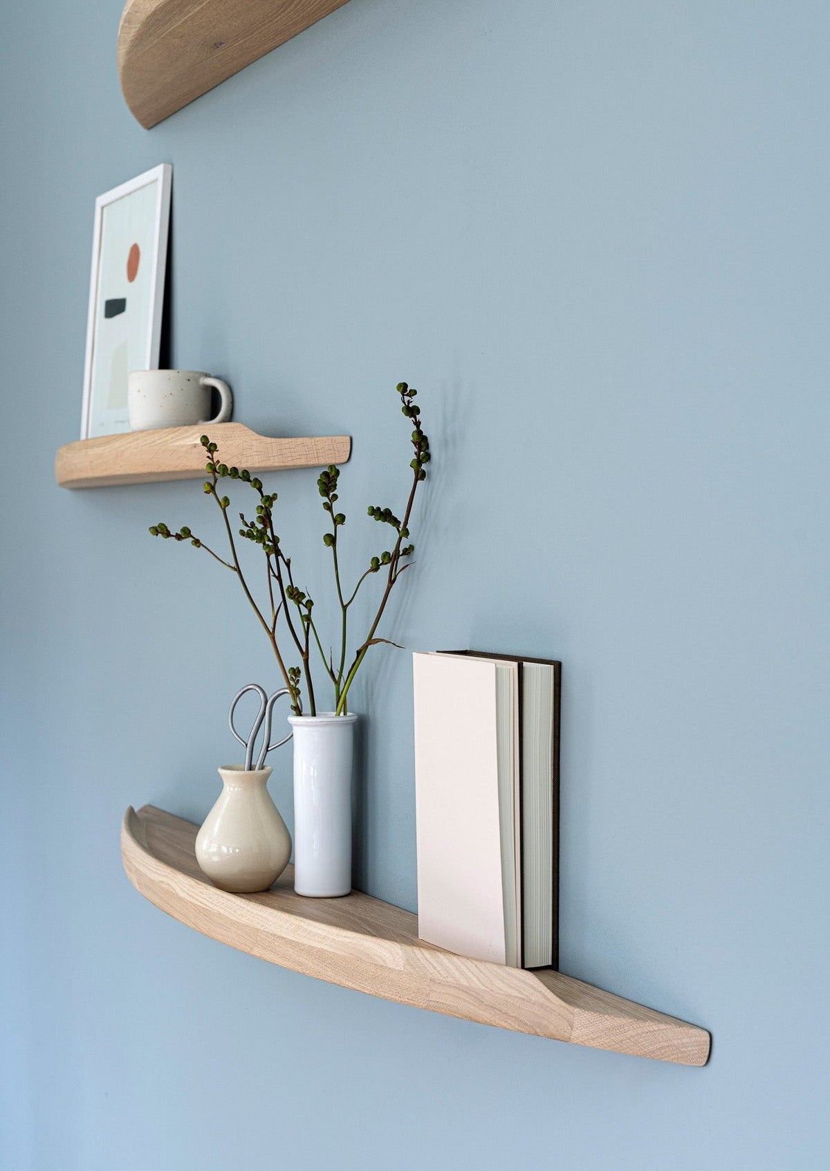 Aakjaer Arcus shelves in white oiled oak holding a print, vases and books on a blue wall