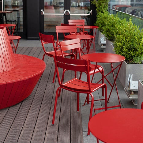 Fermob Bistro Table Red outdoor furniture set with tables and chairs on a wooden deck, large glass windows in the background.